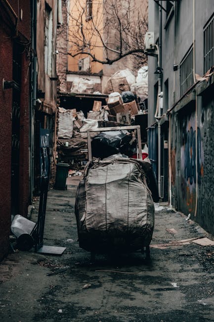 A narrow urban alleyway filled with a substantial accumulation of mixed waste and discarded materials, including a large black plastic bag in the foreground, placed on a wheeled trolley or cart. Behind it, a pile of cardboard boxes, plastic bags, and other refuse dominates the background, stacked against a makeshift barrier or wall. The alley is flanked by tall, weathered buildings, with graffiti visible on the right-hand side wall, and the surfaces appear aged with peeling paint and exposed brick. Bare tree branches extend overhead, adding to the somewhat neglected environment. The ground is uneven, dark, and stained with dirt and debris, reflecting ambient light that highlights the cluttered scene. This scene exemplifies an urgent need for rubbish removal services, such as those provided by Rubbish Clearance Kennington, which specializes in professional waste clearance and independent rubbish collection solutions typical of private disposal in urban contexts.