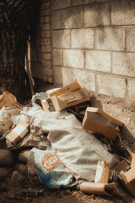 A pile of mixed waste materials sits on the ground adjacent to a brick wall and a tree trunk. The pile includes flattened cardboard boxes with printed labels and logos, crumpled brown paper bags, and large plastic garbage bags filled with unknown contents. Some boxes are partially crumpled or torn, revealing their corrugated texture, while others are intact, showing printed product labels and barcodes. The waste is loosely stacked, with some items leaning against the brick wall, which has a weathered appearance with visible mortar joints and a beige tone. The surrounding environment suggests an outdoor area, possibly a driveway or back alley, with uneven ground composed of dirt and small rocks. The scene appears to be part of a rubbish removal process organized by Rubbish Clearance Kennington, which provides private waste collection services, removing rubbish from such outdoor locations for environmentally responsible disposal.