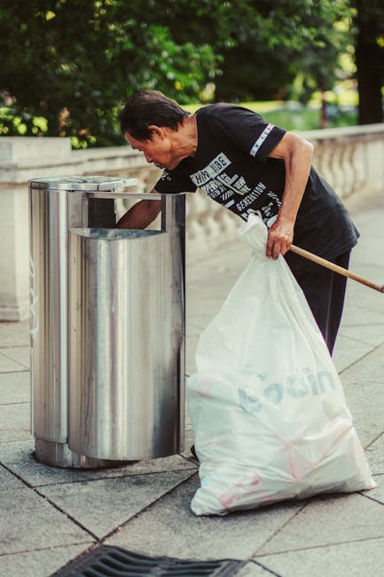 A middle-aged woman with short, dark hair is seen disposing of waste into a cylindrical stainless steel public trash bin with a rounded, open top on a paved outdoor area. She is wearing a black T-shirt with printed text and dark trousers, and is using her right hand to lower a white plastic bag filled with rubbish into the bin while holding a long-handed object, possibly a broom or litter picker, in her left hand. The surrounding environment features a low stone balustrade and lush green foliage, with sunlight filtering through the trees, creating a bright and clean scene. This scene exemplifies urban waste disposal and situational rubbish removal, aligning with private or independent waste management services like those offered by Rubbish Clearance Kennington, especially in the context of alternate rubbish collection options outside local authority provisions.