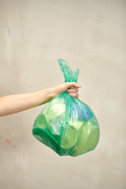 A person's arm is extended from the left side of the image, holding a green plastic rubbish bag filled with waste. The bag appears to be lightweight, slightly crumpled, and tied at the top with a twisted knot. The waste inside the bag includes visible pieces of plastic and other refuse, giving it a semi-transparent appearance that reveals the contents. The background is an off-white, neutral-toned wall with subtle texture, providing a simple backdrop that emphasizes the bag and the arm. The lighting is soft and even, with no harsh shadows, creating a clear and professional visual suitable for a rubbish removal service context. This scene demonstrates the process of discarding waste, aligning with services like private rubbish collection or on-site clearance often provided by companies such as Rubbish Clearance Kennington. The image captures a moment of waste disposal, highlighting the importance of proper rubbish handling and the role of independent waste removal services in managing household or commercial waste efficiently.