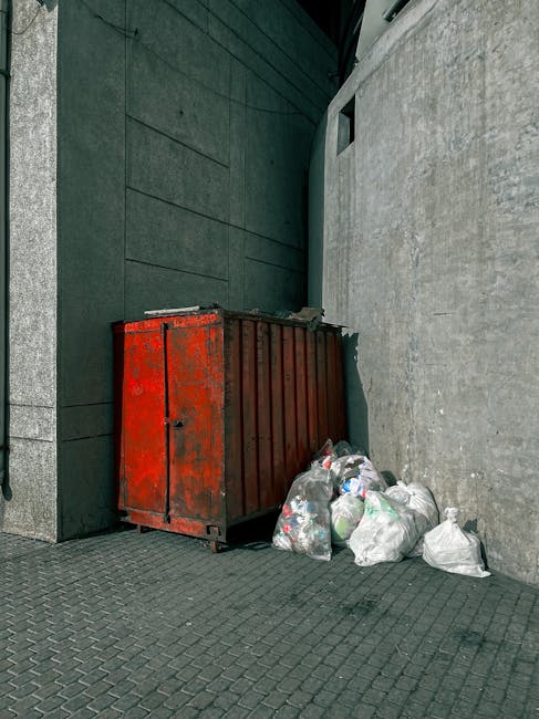 A large, rusted metal skip with a red finish, positioned against a dark green wall on the left and a light grey concrete wall on the right on a paved urban sidewalk. The skip contains some debris visible along its open top edge, and several white plastic bags filled with waste are placed on the ground around its base, leaning against the right side of the skip. The scene appears to be an outdoor area designated for waste accumulation, possibly in an alleyway or behind commercial premises, with minimal natural lighting and a muted overall atmosphere. This setup is indicative of private rubbish collection services, such as those offered by Rubbish Clearance Kennington, supporting alternative waste management solutions outside of municipal disposal channels. The environment emphasizes the functional and utilitarian nature of on-site waste storage, prepared for removal or clearance.