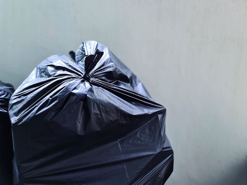 A pile of discarded cardboard boxes, some flattened and others partially intact, rests against a weathered brick wall outdoors. The boxes, varying in size and mainly brown with printed labels and barcodes, are interspersed with crumpled brown paper and white packaging materials. A large, textured fabric sack or bag, possibly used for waste collection, is partially covering some boxes and is tied at the top. Surrounding the pile are small loose stones and debris on the ground, with a tree trunk visible to the left side of the image. The scene suggests an area designated for waste or rubbish accumulation, possibly as part of an on-site clearance or private waste disposal process typically managed by local rubbish removal services, like Rubbish Clearance Kennington, to facilitate efficient rubbish collection and site cleanup.