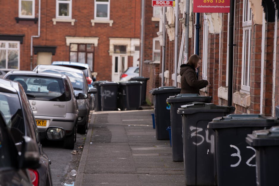 A middle-aged man with dark hair, wearing a black T-shirt with graphic white text on the front, is seen disposing of rubbish into a dedicated metal waste bin on a city sidewalk. He is holding a large white plastic bag filled with waste in his left hand, while his right hand is leaning into the bin to deposit the trash. The metal bin has a sleek, cylindrical shape with a shiny, reflective surface, featuring an opening at the top for waste disposal. The scene is set outdoors in a well-maintained urban environment, with a concrete pavement beneath the man’s feet and a decorative stone balustrade visible in the background. Surrounding the area are lush green trees and foliage, suggesting a park-like setting. The lighting is natural, with soft daylight filtering through the trees, providing a clear view of the waste collection process, which may reflect the type of independent or alternative waste handling service provided by Rubbish Clearance Kennington in managing rubbish removal tasks on-site or in public spaces.