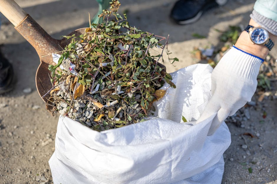 A person wearing a white glove and a wristwatch is holding a white heavy-duty rubbish bag filled with garden debris, including small branches, leaves, and twigs, while standing on a gravel surface. A small, rusty shovel with a wooden handle is partially inserted into the bag, pressing down the contents. The environment appears to be an outdoor area, possibly a garden or street, with natural sunlight casting shadows and highlighting the textured materials. This scene depicts the transfer of garden waste for removal, demonstrating a typical activity associated with private rubbish collection or clearance services like those offered by Rubbish Clearance Kennington, facilitating on-site or alternative waste handling solutions. The visible details of the debris and tools emphasize professional rubbish removal processes aimed at clearing outdoor spaces by collecting biodegradable waste efficiently.
