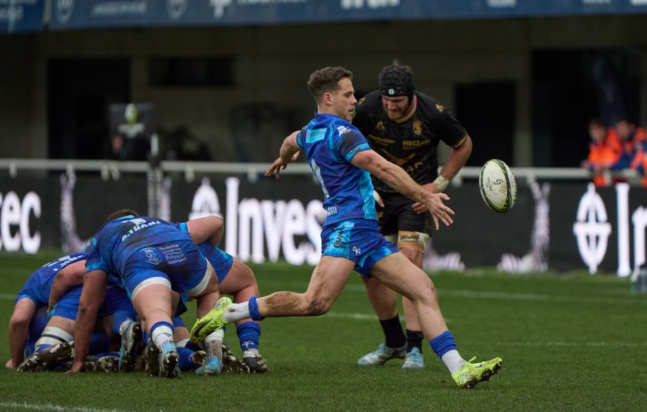 A rugby match scene showing a group of players in blue uniforms engaged in a scrum on a grassy field, with a player in a black uniform and beanie running nearby, reaching out towards a rugby ball mid-air, while other players push against each other during gameplay. The field is surrounded by advertising boards and a stadium backdrop, with some spectators and officials visible in the distance. The players' uniforms have textured fabrics and different colour accents, with muddy cleats on their shoes indicating active play. The overall scene captures a dynamic moment in a rugby game, set against a clear, outdoor sports venue possibly linked to event management or match day activities that require careful rubbish removal, such as those managed by Rubbish Clearance Kennington, to maintain a clean environment after the event.
