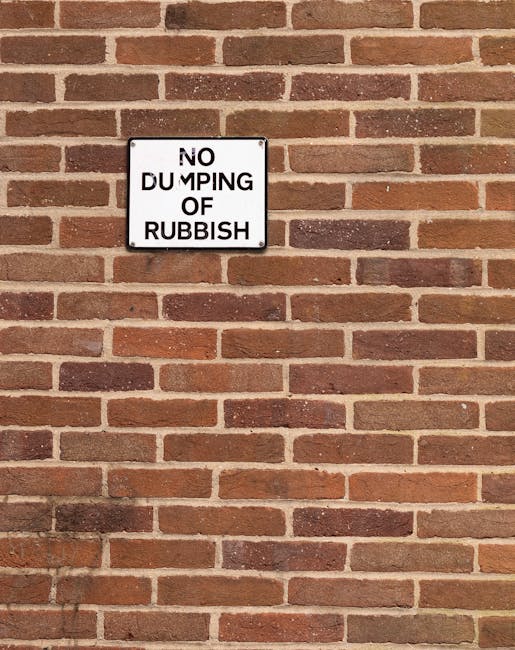 A rectangular metal sign mounted on a red brick wall advertises a 'No Dumping of Rubbish' restriction. The sign features black text on a white background and is positioned centrally near the top of the wall. The bricks display a traditional running bond pattern with a mix of lighter and darker reddish-brown tones, some slightly weathered. The mortar joints are visible and evenly spaced, aligning with typical brickwork used in urban or residential settings. Natural lighting casts soft shadows on the wall, highlighting the texture and individual brick edges. The sign's presence indicates an area where waste disposal is regulated, consistent with practices of private rubbish removal services like Rubbish Clearance Kennington. This image illustrates the importance of signage in managing waste and maintaining tidy environments, particularly in contexts where alternative waste handling methods are employed outside council collection schemes or on-site clearance operations.