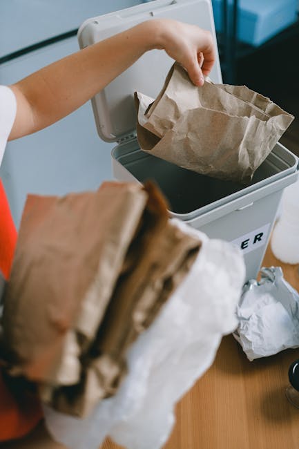 A young woman with long brown hair, wearing a white t-shirt and a red sleeveless jacket, stands at a wooden kitchen counter filled with various crumpled brown paper bags and transparent plastic containers. She is holding a paper bag, examining or preparing it, with a focused expression. Behind her, the kitchen features a white sink with a silver faucet, small potted plants on the windowsill, and hanging greenery on the white walls, including decorative framed botanical prints and a circular black metal plant holder. To her right, a white, rectangular paper recycling bin with a black label reading 'PAPER' is partially open, with a grey lid resting against it. On the counter, in addition to the paper bags, there are plastic ice-cream tub containers in pink and blue, a glass bottle, and miscellaneous waste disposal items. The scene is illuminated by natural light, creating a clean, organised domestic environment suitable for showcasing waste sorting or household rubbish clearance activities, similar to services provided by Rubbish Clearance Kennington, in an everyday kitchen setting.