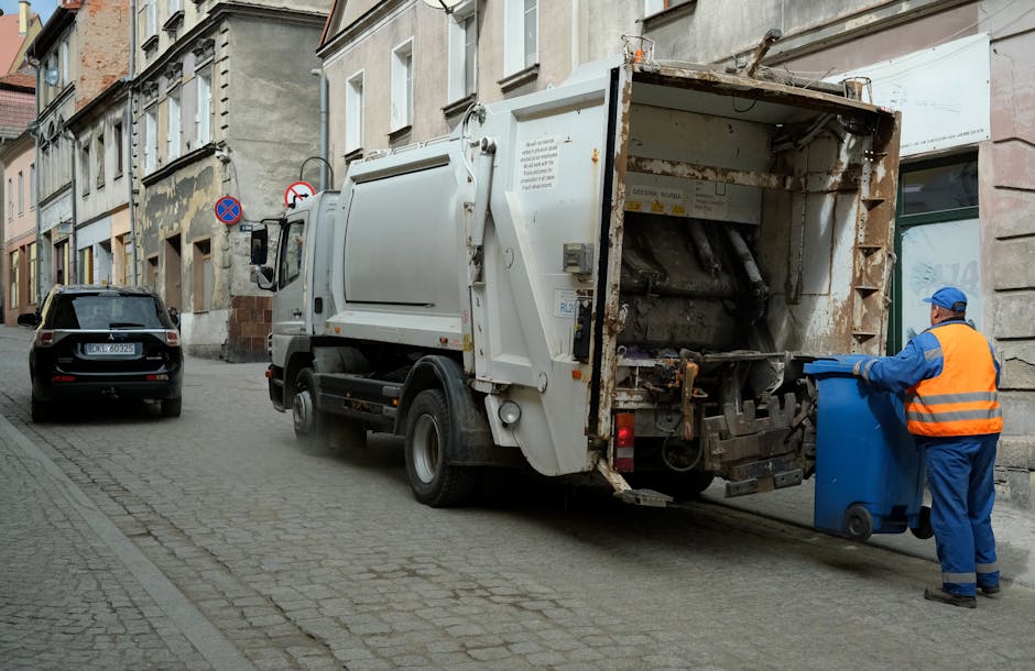 A waste collection vehicle parked on a narrow urban street in front of old multi-story buildings with weathered facades. The truck is primarily white with some rust and dirt visible on its rear loading section, which is open and partially empty, designed for the collection of bulky waste or rubbish. A worker wearing a blue uniform and high-visibility orange vest is positioned at the rear, holding a blue wheeled bin and preparing to empty it into the truck. The street is paved with cobblestones and shows typical city congestion with another parked vehicle, a small black SUV, seen on the left side of the image. Environmental lighting suggests daylight, with diffused natural light illuminating the scene, appropriate for documenting a waste removal service focused on rubbish clearance or on-site waste disposal, similar to private waste collection activities undertaken by companies like Rubbish Clearance Kennington. Environmental context shows an urban setting where independent rubbish collection services might operate outside of municipal disposal systems, emphasizing professional rubbish removal in a neighbourhood setting.