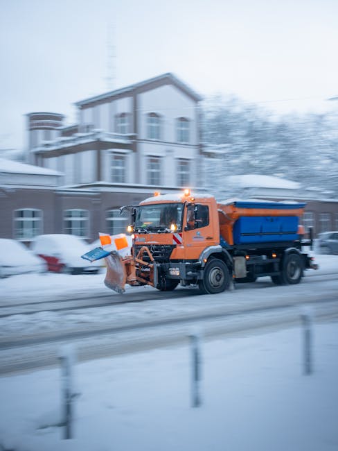 An orange waste collection truck equipped with a plow attachment is actively clearing snow and debris from a street in front of a residential building during winter. The truck features a blue skip at the rear, used for collecting and transporting rubbish, and has flashing amber lights on the roof for safety. The background shows a multi-storey house with white walls, dark window frames, and a snow-covered roof, while parked cars on the street are partially visible beneath a layer of snow. The scene is characterized by overcast, grey lighting typical of winter weather, and the snow on the ground appears compacted and wet. The presence of the rubbish vehicle indicates an on-site or private waste removal service, supporting alternative rubbish collection methods outside of local authority services. The overall environment depicts a typical urban residential area undergoing street cleaning and rubbish clearance during cold weather, with Rubbish Clearance Kennington specializing in such services.