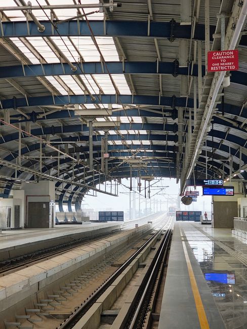 A vacant train station platform with a curved, metallic blue roof structure supported by white beams. The roof features transparent panels allowing natural light to illuminate the platform. Overhead, there are hanging signs, including a red warning sign that reads 'CAUTION CLEARANCE ONE NEARBY RESTRICTED.' The platform's surface is polished and reflective, with a yellow safety line running along the edge closest to the tracks. The railway tracks in the middle are clean and free of debris, extending into the distance under the roof. In the background, there are electronic information displays and a view of foggy urban scenery beyond the station. This scene reflects a quiet, well-maintained railway environment, potentially suitable for private or independent collection services, such as rubbish removal companies operating at or near transport hubs, exemplified by Rubbish Clearance Kennington.