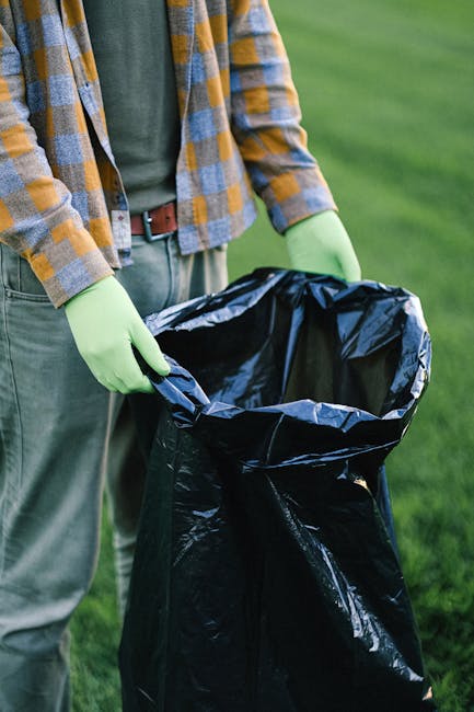 The image depicts a person outdoors, standing on a grassy area, holding a large black rubbish bag made of shiny, durable plastic. The individual is dressed casually in a plaid shirt featuring shades of yellow, blue, and grey, over a dark green T-shirt, paired with grey trousers and a brown belt. They are wearing light green rubber gloves, suggesting an active engagement in waste collection or disposal. The person's hands are gripping the top of the rubbish bag, which is open, ready for filling with waste. The background shows well-maintained, vibrant green grass, indicating a park, garden, or open outdoor space. The scene emphasizes controlled waste collection, possibly as part of an attempt to manage rubbish independently, aligning with the context of alternative rubbish removal services. The lighting appears natural, highlighting the textures of clothing, gloves, and the reflective surface of the plastic rubbish bag, ensuring all elements are clearly visible and detailed for accessibility purposes.