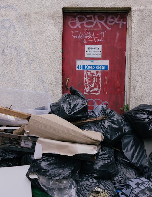 A close-up view of a weathered red metal door mounted in a beige brick or concrete wall, with graffiti tags in white and pink spray paint on its surface. The door features a small rectangular sign indicating 'NO MONEY' and 'Keep Clear' in blue. Surrounding the door, there is a significant accumulation of discarded rubbish, including black garbage bags, flattened cardboard boxes, and loose debris, piled up at the base and partially leaning against the door. The rubbish appears to contain mixed household waste, and the bags are tightly knotted, with some showing signs of tearing. The environment suggests an urban back alley or service area, with the rubbish indicating a private waste collection or on-site clearance scenario, highlighting the need for professional rubbish removal services such as those offered by Rubbish Clearance Kennington. The lighting is natural, with soft shadows cast across the scene, emphasizing the clutter and the aged condition of the door and surrounding wall.