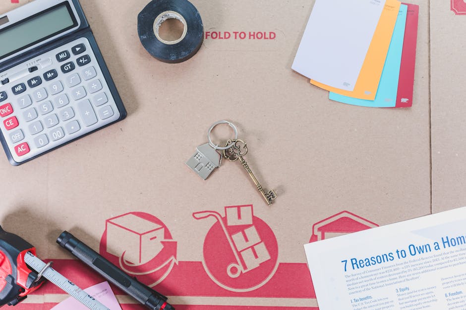 A flat-lay photograph displays various office and packing items arranged on a light brown cardboard surface, which features red printed symbols related to logistics and waste handling, including a cube, a hand truck, and an arrow circling a box. In the center, a silver keyring with a house-shaped and a rectangular tag is clearly visible alongside an old-fashioned, ornate brass key. On the top left, there is a partial view of a black calculator with white and pink buttons facing upwards. To the top right, a small stack of unclipped pastel-colored paper sheets—white, yellow, blue, and pink—are neatly arranged. Near the top center, a roll of black duct tape with a cardboard core rests on the surface, and the words 'FOLD TO HOLD' appear in red text around it. A black pen with a textured grip and metal clip is situated towards the bottom left, next to a silver and pink ruler. In the bottom right corner, a white sheet of paper printed with blue text titled '7 Reasons to Own a Home' is partially visible. The arrangement and objects evoke themes of moving, organization, and waste management, aligning subtly with the context of rubbish removal services. The lighting is even and natural, emphasizing clear visibility of each item without harsh shadows or reflections.